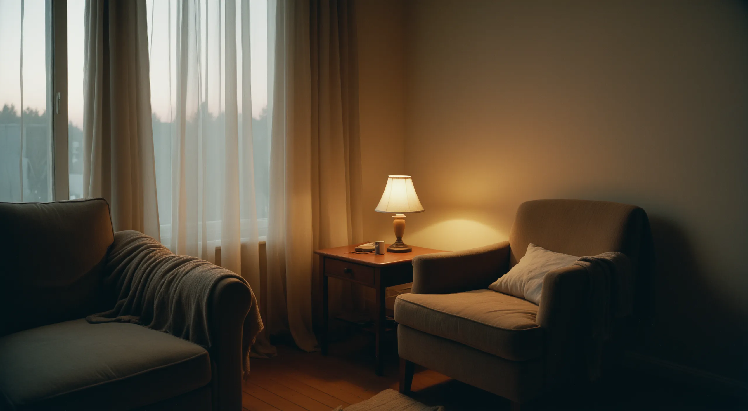 A dim, calm room at dusk lit by a single warm lamp, sheer curtains over the window
